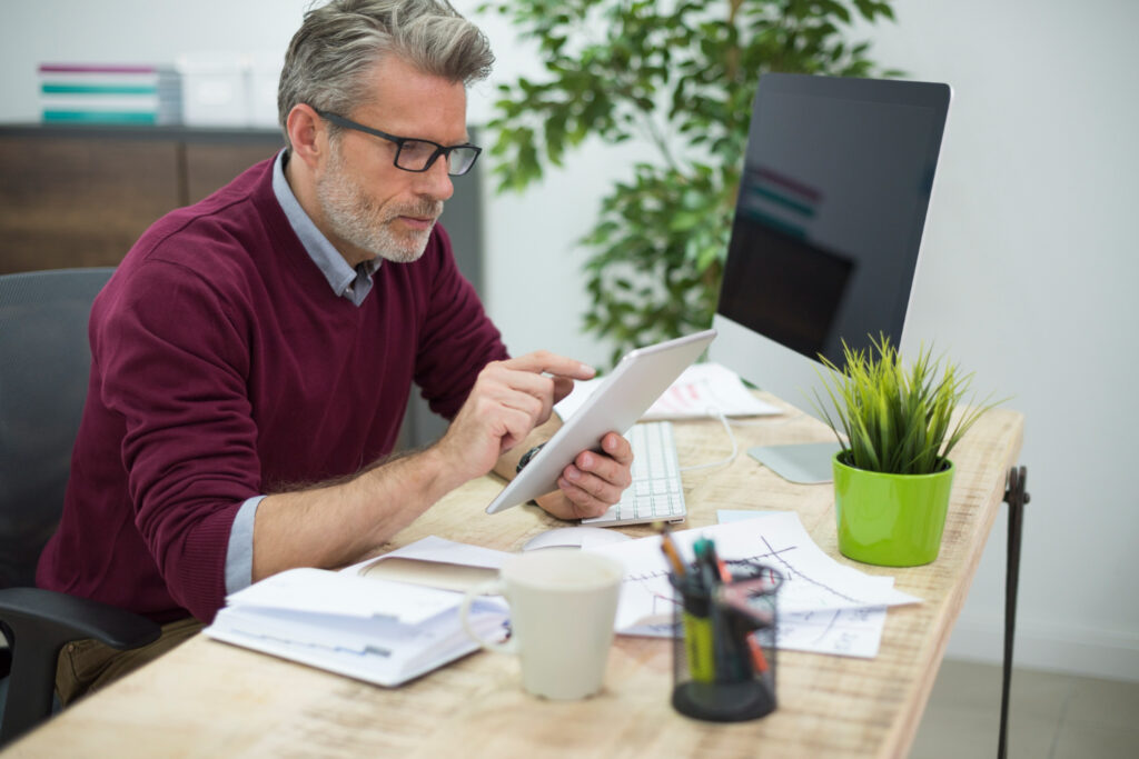 A man with glasses works at a desk, using a tablet. Papers, a mug, pens, a plant, and a computer monitor are on the desk. A leafy plant is in the background. | Betechit.com