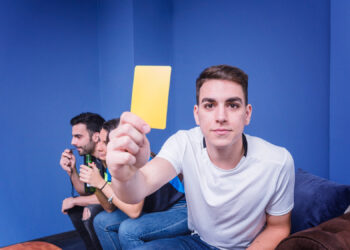 A young man in a white shirt sits on a couch, holding a yellow card toward the camera. Two other men sit beside him in the background, appearing to watch something intently. The room has blue walls. | Betechit.com