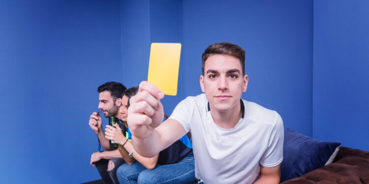 A young man in a white shirt sits on a couch, holding a yellow card toward the camera. Two other men sit beside him in the background, appearing to watch something intently. The room has blue walls. | Betechit.com