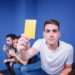 A young man in a white shirt sits on a couch, holding a yellow card toward the camera. Two other men sit beside him in the background, appearing to watch something intently. The room has blue walls. | Betechit.com