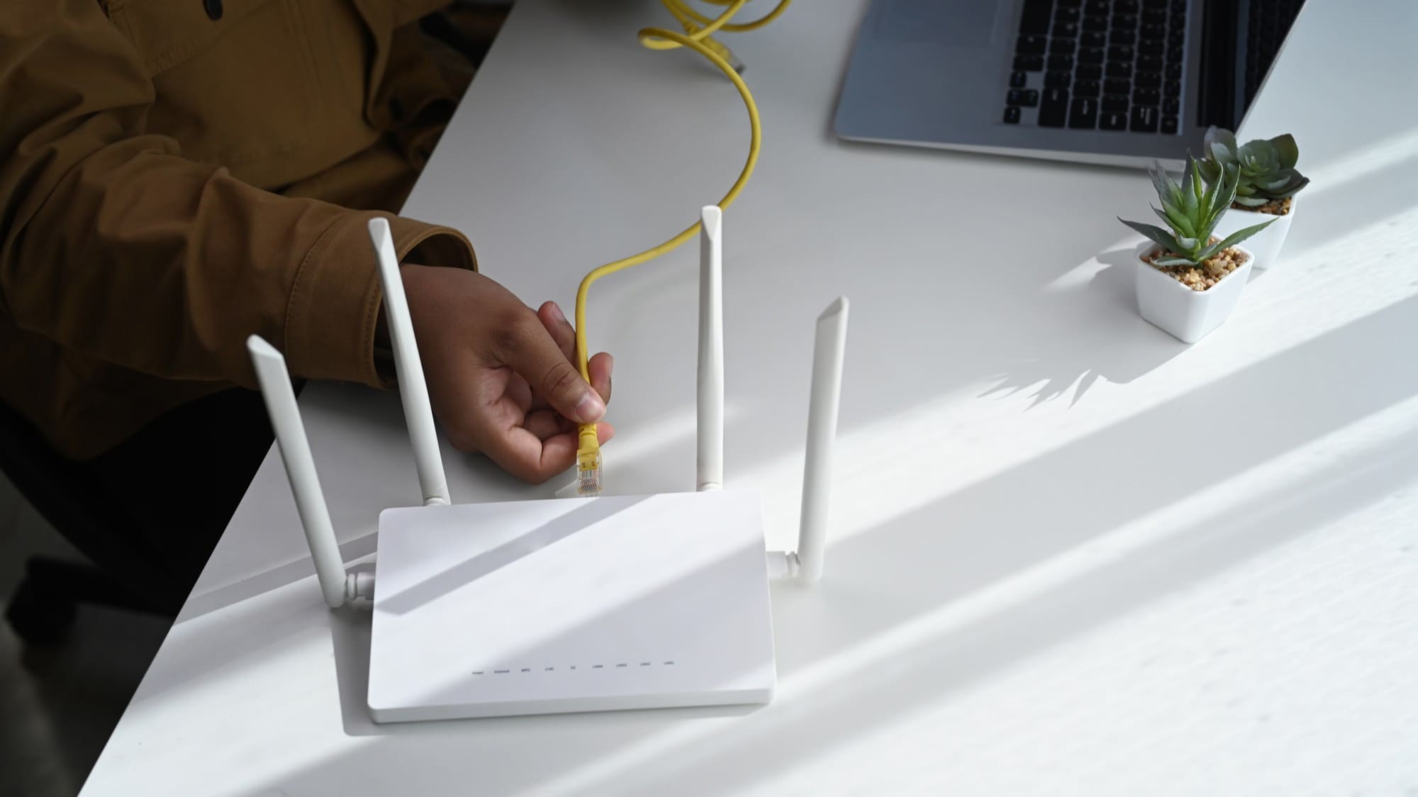 A person connects a yellow Ethernet cable to a white wireless router on a white desk, with a laptop and a small potted plant nearby. | BeTechIT