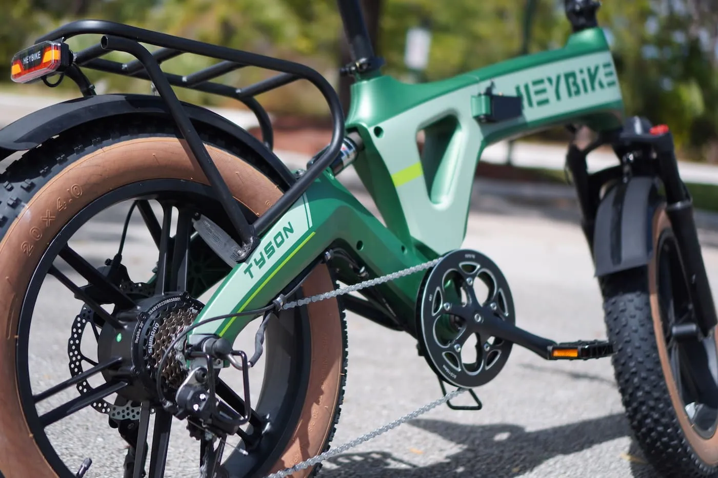 Close-up of a green Heybike Tyson electric bicycle, highlighting the rear wheel, gears, pedals, brown tires, and rear cargo rack—perfect for sports enthusiasts—set against a blurred outdoor background. | BeTechIT