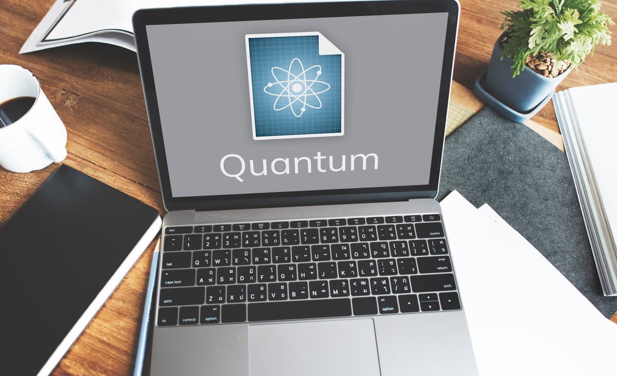 A laptop displaying a screen with a document icon featuring an atomic symbol and the word "Quantum." The laptop is on a wooden desk with notebooks, papers, a pen, and a cup of coffee nearby. | Betechit.com