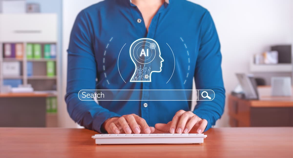 A person in a blue shirt types on a keyboard at a desk, with a digital illustration of a human head containing "AI" and a search bar overlayed in front. The background shows a blurred office setting. | Betechit.com