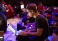 A group of people sitting on bean bags, working on laptops in a dimly lit room. The focus is on a person in glasses typing on a laptop, with others in the background also using computers. | Betechit.com