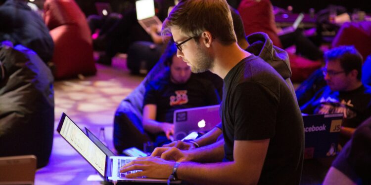 A group of people sitting on bean bags, working on laptops in a dimly lit room. The focus is on a person in glasses typing on a laptop, with others in the background also using computers. | Betechit.com