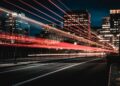 A city street at night with light trails from passing vehicles, surrounded by tall illuminated buildings. The sky is dark, highlighting the vibrant energy of urban life. | Betechit.com