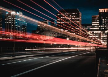 A city street at night with light trails from passing vehicles, surrounded by tall illuminated buildings. The sky is dark, highlighting the vibrant energy of urban life. | Betechit.com