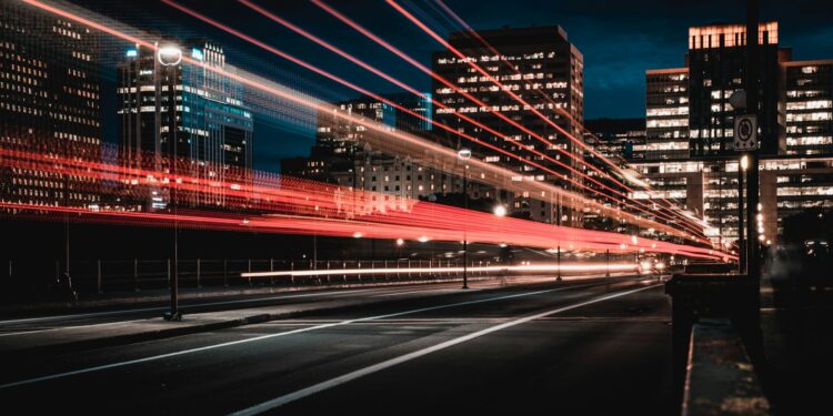 A city street at night with light trails from passing vehicles, surrounded by tall illuminated buildings. The sky is dark, highlighting the vibrant energy of urban life. | Betechit.com